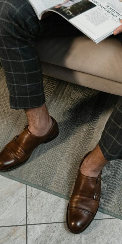 Well-dressed man in brown leather shoes and checkered pants reading a magazine indoors.